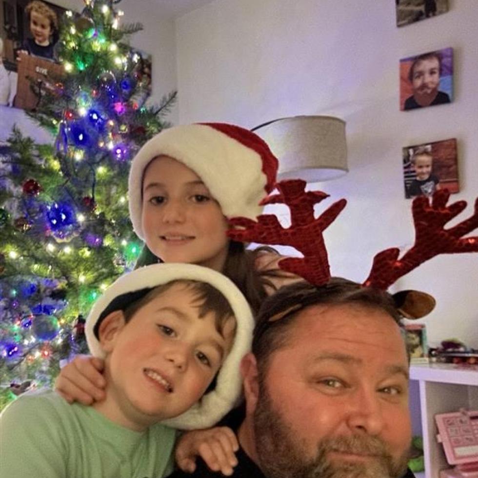 Jeff Davis with his two children: Zoe (9) and Austin (7) wearing red Christmas hats next to a decorated Christmas Tree