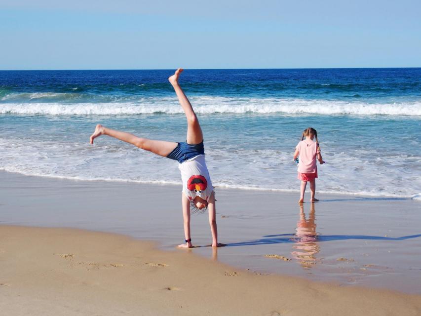 Two kids at the beach, one of them in the water, the other doing a cartwheel