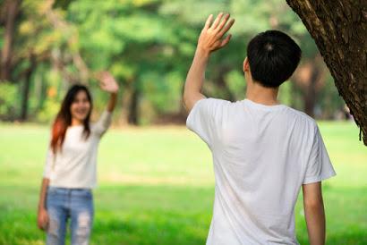 Two teenagers in a park waving at each other from six feet apart in order to practice social distancing.