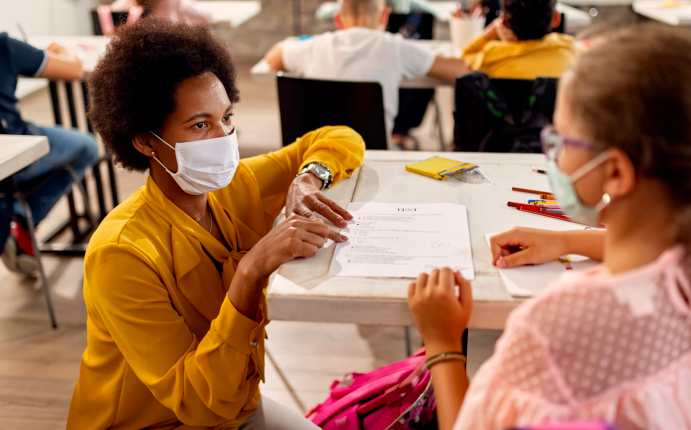 Teacher, wearing a mask, crouching down next to student's desk who is also wearing a mask.