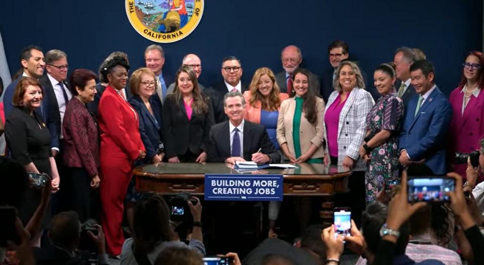 Governor Newsom sitting at a desk signing legislation with a large group of people
